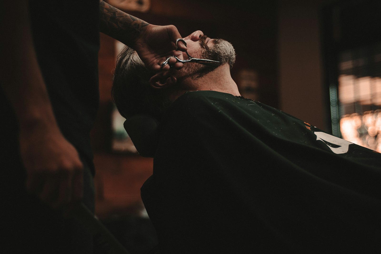 A man in a barber's chair having his beard shaped by a barber